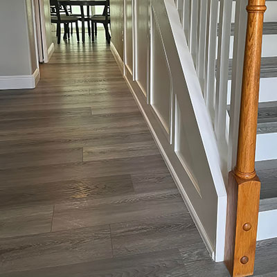 A hallway with gray wood flooring, white wainscoting on the wall along a staircase with a wooden banister, and a dining table with chairs visible in the background.