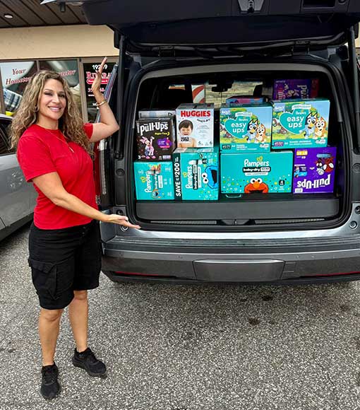 A woman in a red shirt and black shorts stands next to an open car trunk filled with boxes of diapers and pull-ups, smiling and gesturing toward the donation.