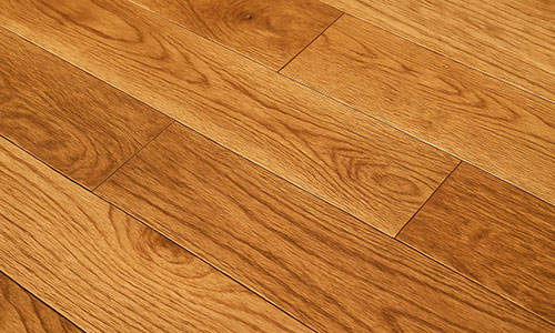 Close-up view of wooden floorboards with a natural grain pattern and a warm, honey-brown finish. The planks are neatly aligned, showcasing the texture and color variations of the wood.