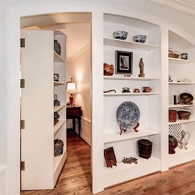 A white built-in bookshelf with various decorative items partially opens to reveal a hidden doorway leading to a warmly lit hallway with a wooden floor and a small lamp on a table.