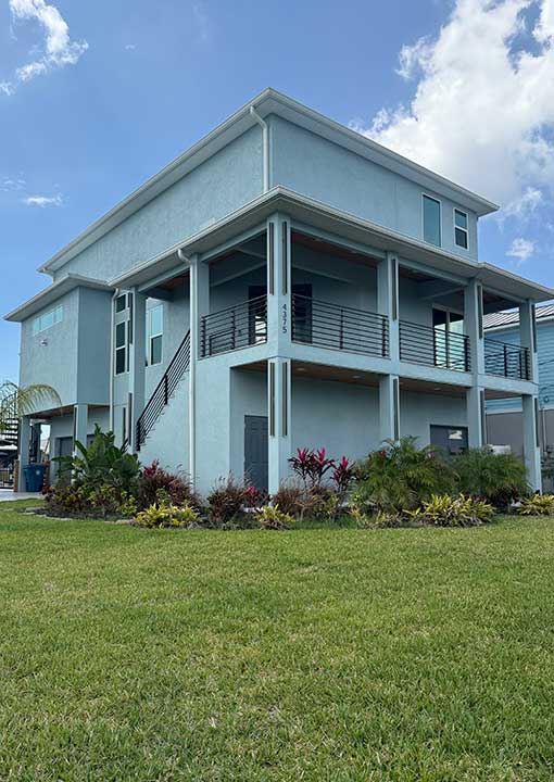 A modern two-story house with light blue exterior, large wraparound balcony, and metal railings, surrounded by green lawn and landscaped plants under a partly cloudy sky.