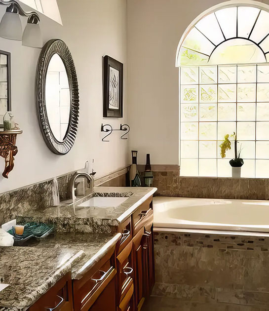 A bathroom with a marble countertop, wooden cabinets, a round mirror, wall-mounted lights, and a bathtub beneath a large arched window with frosted glass blocks and a potted orchid.