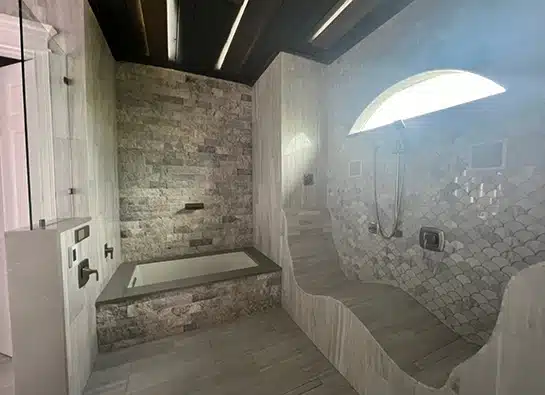 Modern bathroom with stone tile walls, a built-in bathtub, a curved tiled bench, a wall-mounted shower, and a large arched frosted window letting in natural light.