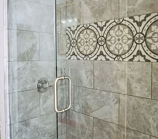 Glass shower door with stainless steel handle, set in a bathroom featuring large gray marble tiles and a horizontal accent strip of decorative patterned tiles in black and white.