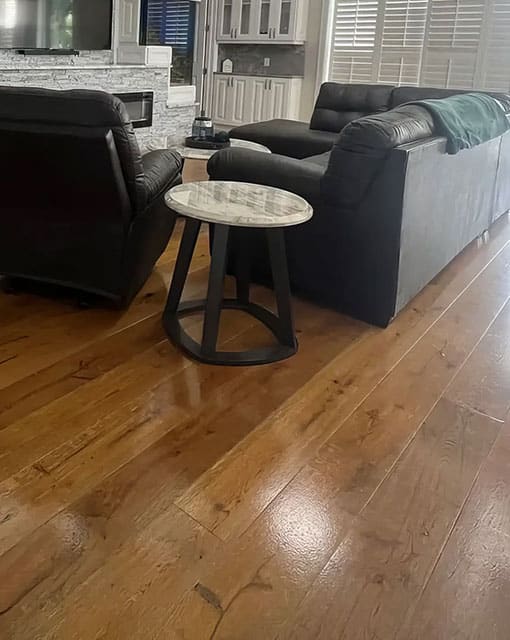 A living room with wooden floors, a round marble-top side table, two black leather sofas, and a blanket draped over one sofa. The background shows a kitchen area with white cabinets and large windows.