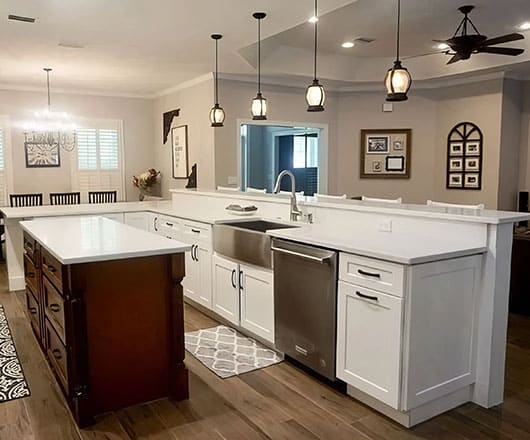 Modern kitchen with white cabinets, stainless steel appliances, farmhouse sink, large island with dark wood base, hanging pendant lights, and adjacent dining area with wall decor and wood flooring.