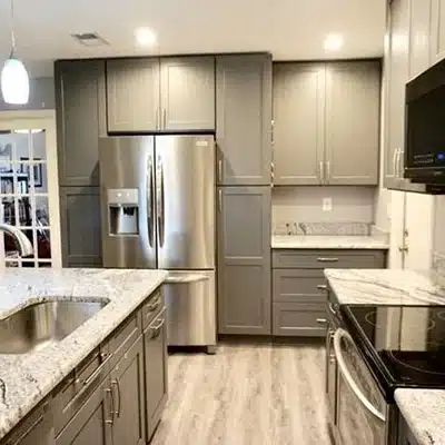 Modern kitchen with gray cabinets, stainless steel appliances, marble countertops, and wood-style flooring. The layout includes an island with a sink, built-in refrigerator, oven, and microwave. Bright lighting overhead.