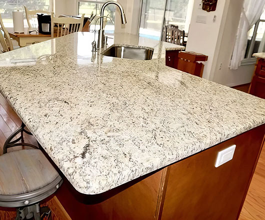 A kitchen island with a polished light-colored granite countertop, a built-in sink with a modern faucet, and wooden cabinets below, situated in a bright, sunlit kitchen.
