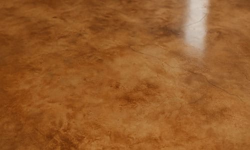 Smooth, polished concrete floor with a mottled brown and amber finish, featuring a subtle sheen and some faint cracks and light reflections.