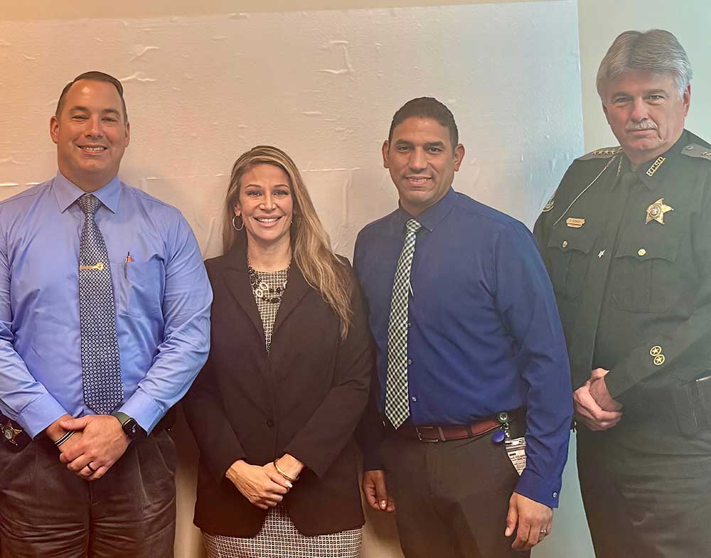 Four adults pose for a group photo against a light wall. Three are wearing blue shirts and business attire, and one is in a dark green sheriff's uniform with a badge. All are facing the camera and smiling.