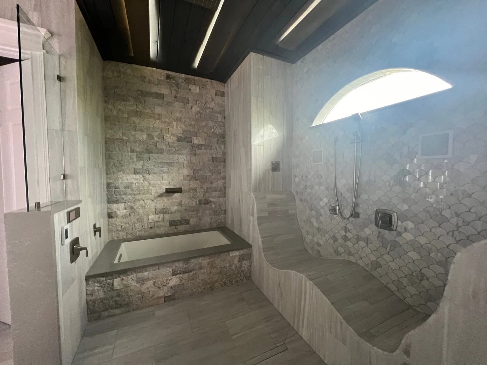 A modern bathroom with stone tile walls, a built-in rectangular bathtub, a curving tiled shower bench, and a large arched window above the shower area. The ceiling is dark with recessed linear lighting.