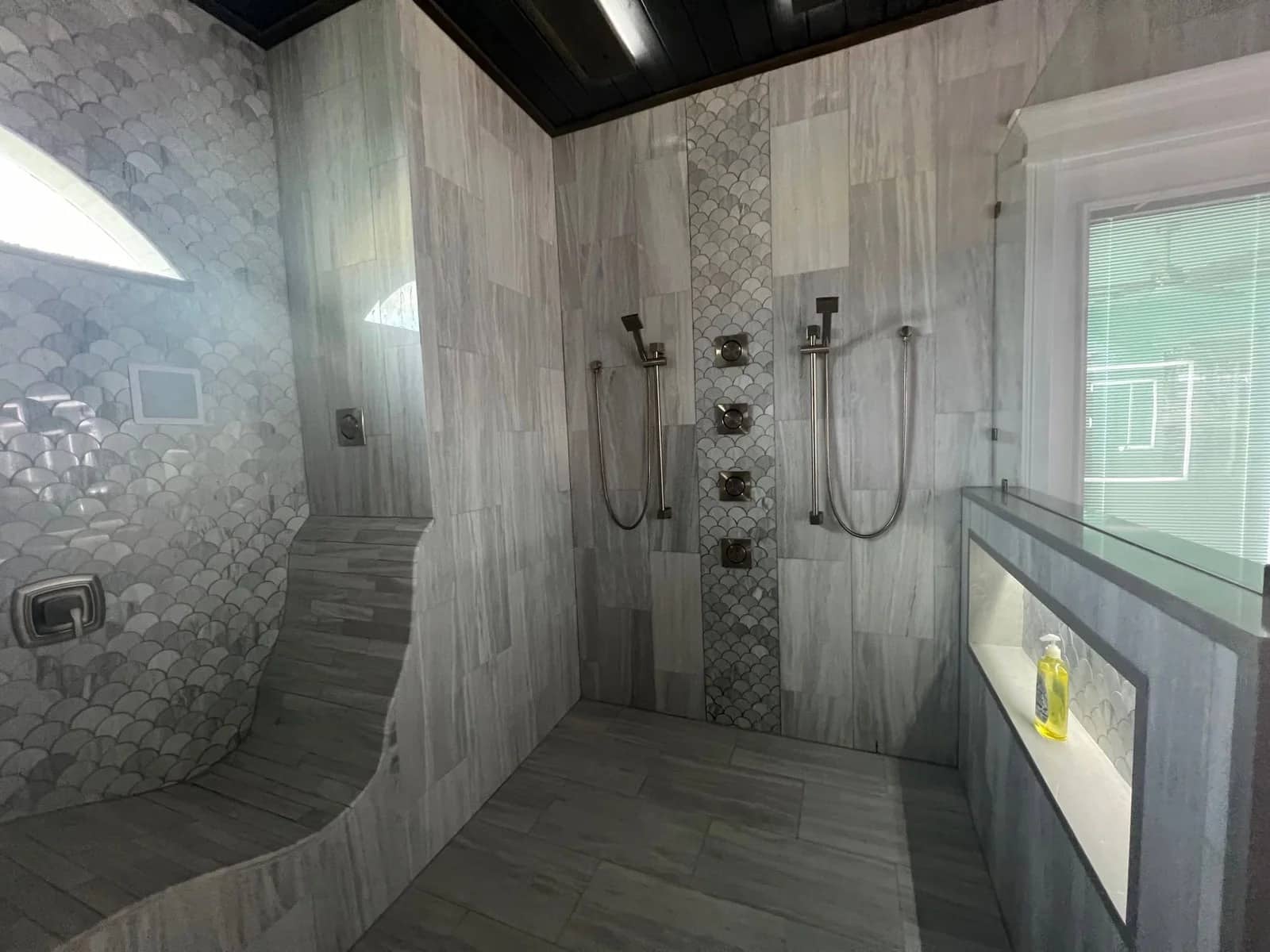 Modern shower with light gray tile, dual showerheads, multiple wall jets, a built-in bench with fish-scale tile design, and a recessed shelf holding a clear bottle of yellow liquid. Natural light enters through a window.