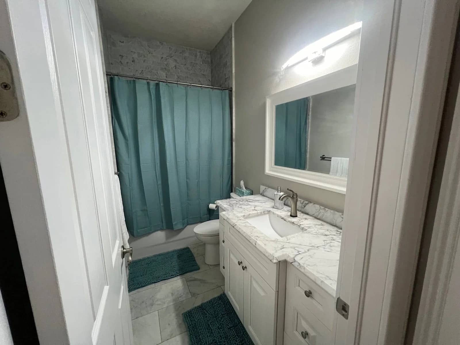 A small bathroom with white cabinets and marble countertops, a sink, mirror, toilet, and a bathtub with a teal shower curtain. There are matching teal rugs on the white tile floor. The walls are painted light gray.