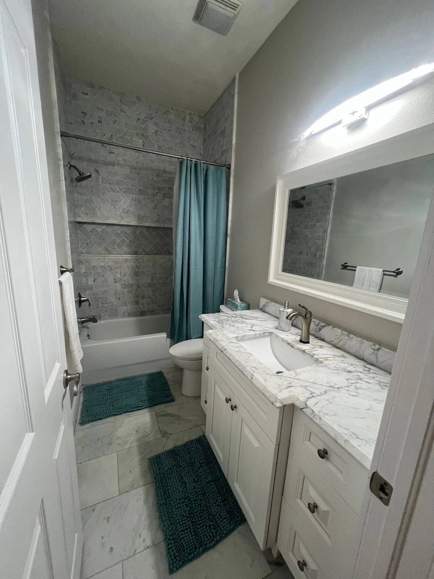 A modern bathroom with a marble countertop vanity, white cabinets, a large mirror, bathtub with a teal shower curtain, and matching teal bath mats on a light tile floor. Towels hang on the wall next to the tub.