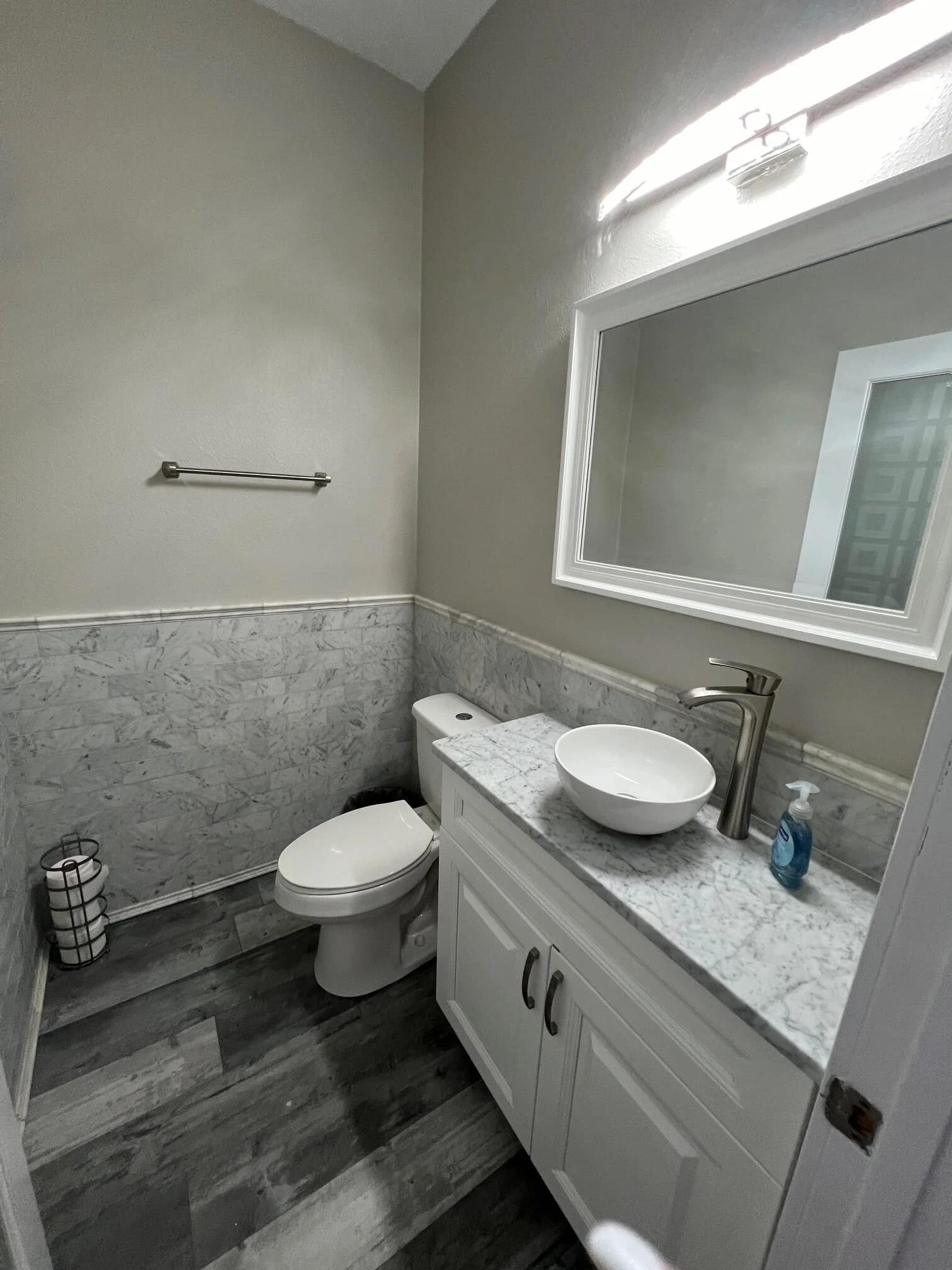 A modern bathroom with gray marble flooring, a white toilet, a white vanity with a vessel sink, a large mirror above the sink, light gray walls, and a hand soap bottle on the counter.