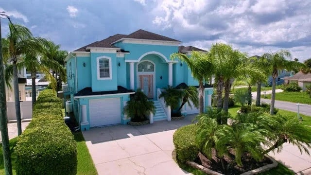 A large, two-story turquoise house with white trim, surrounded by palm trees and greenery, sits under a partly cloudy sky. The driveway leads to a garage, and steps lead to the front door.