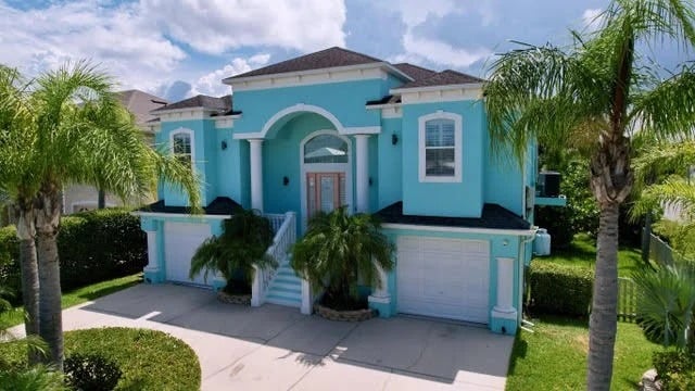 A large, two-story turquoise house with white trim, tall arched entryway, double doors, two garages, palm trees, and well-manicured landscaping under a partly cloudy sky.