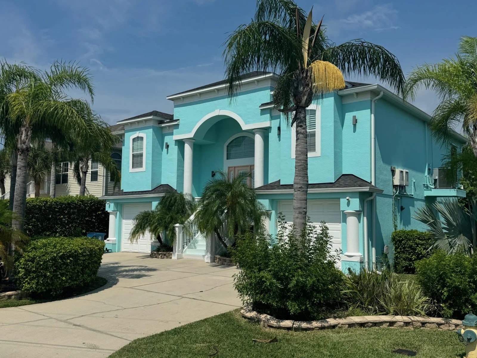 A two-story turquoise house with white trim, surrounded by palm trees and lush greenery, featuring three garage doors, arched entryway, and a curved driveway under a partly cloudy sky.