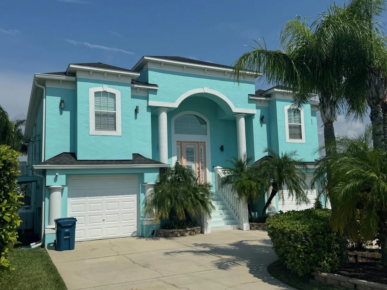 A two-story turquoise house with white trim and double garages, surrounded by palm trees and greenery, under a clear blue sky. White stairs lead to the main entrance with tall columns.