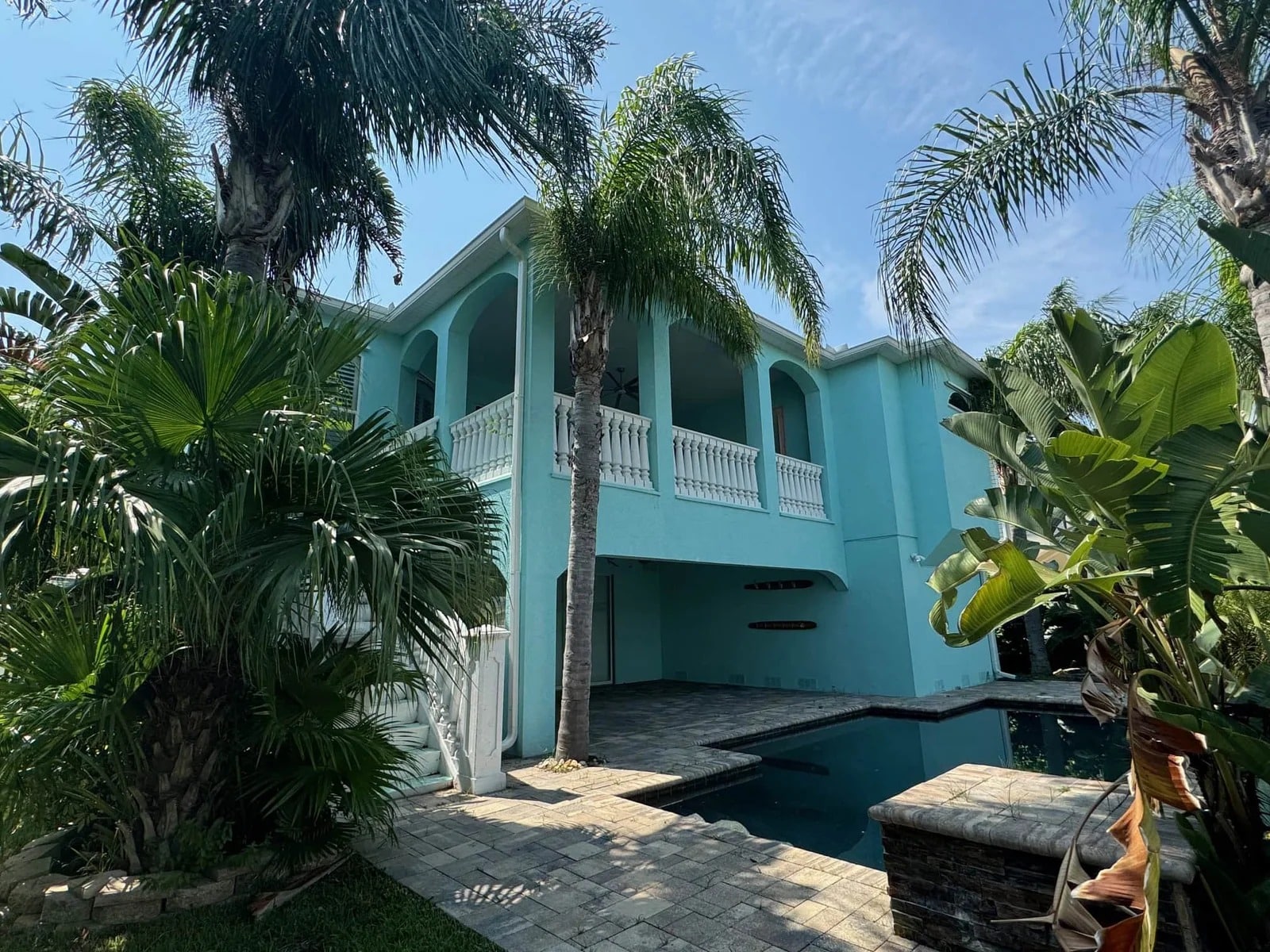 A two-story turquoise house with white railings and columns, surrounded by tall palm trees and lush greenery, features a paved patio area and a small swimming pool under a clear blue sky.