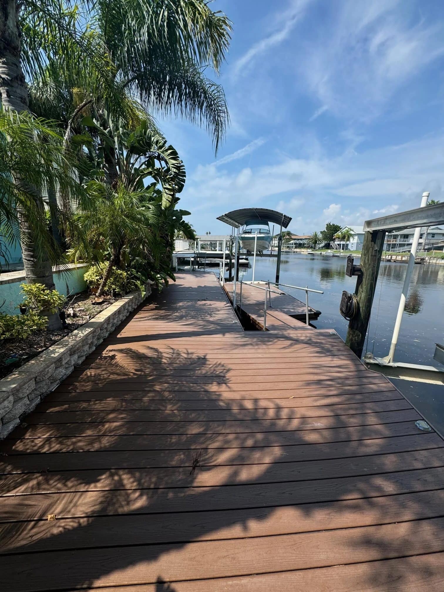 A wooden dock lined with palm trees extends alongside calm water in a canal, with a boat lift and residential houses visible in the background on a sunny day.