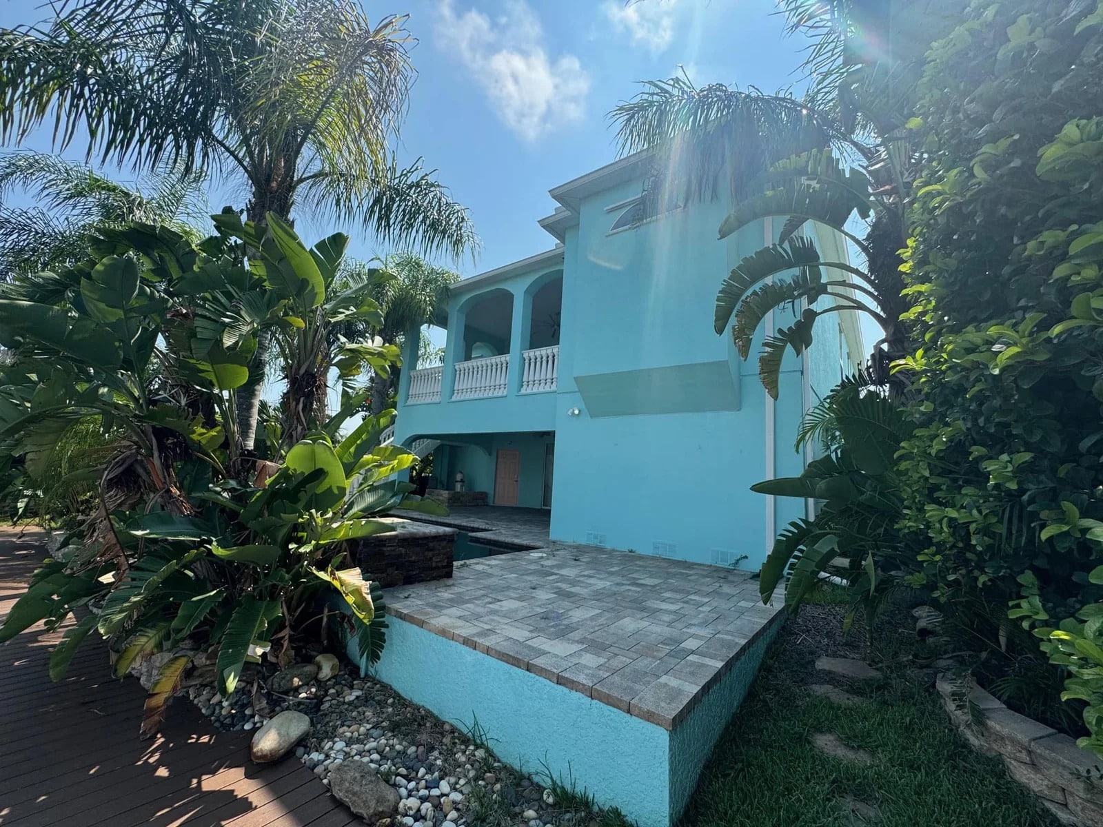 A two-story light blue house with a covered balcony, surrounded by lush tropical plants and palm trees, under a bright blue sky with scattered clouds. The foreground features a paved patio area.