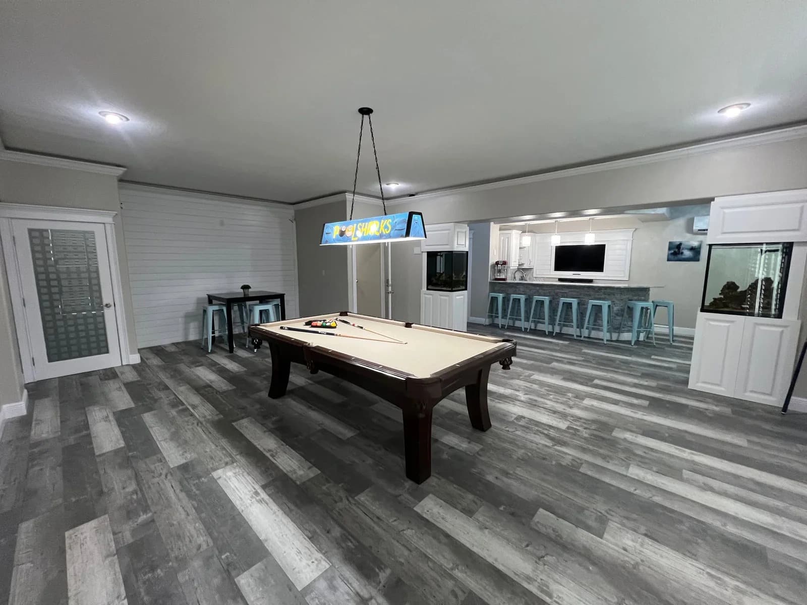 A modern game room with a pool table at the center, hanging light fixture above, gray wood flooring, and an open kitchen area with barstools and aquariums in the background.