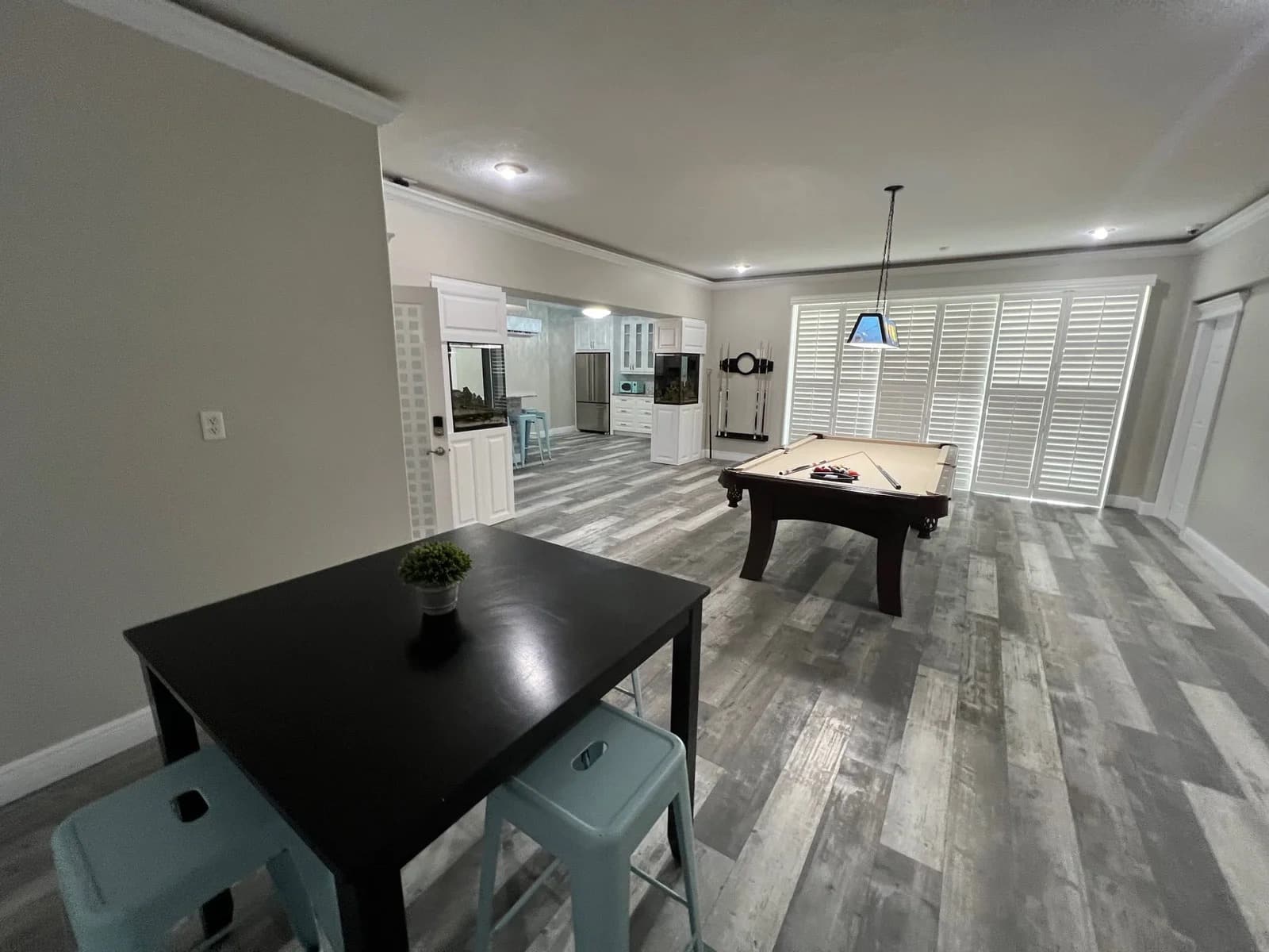 A modern game room with gray wood flooring, a square black table with four stools, a small plant, and a pool table under a hanging light. Large windows with white shutters let in natural light.