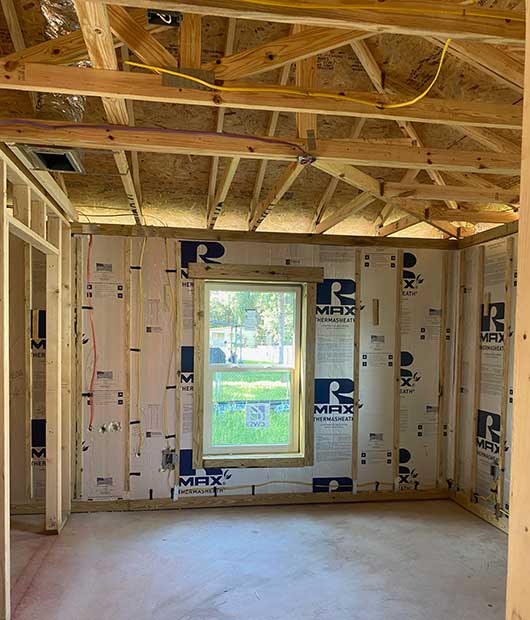 A room under construction with exposed wooden framing, insulation panels on the walls, a window in the center letting in daylight, visible wiring, and an unfinished concrete floor.