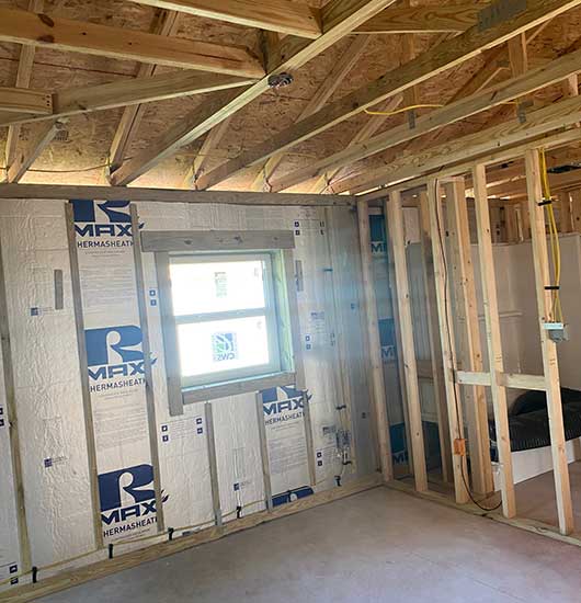 A room under construction shows exposed wooden framing and ceiling beams, a window, insulation panels on the walls, visible wiring, and unfinished flooring. Natural light enters through the window.