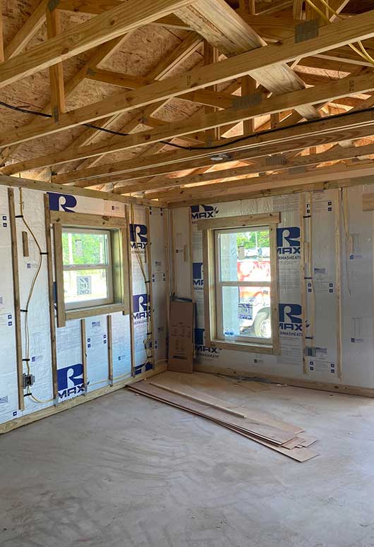 Interior of a house under construction, showing exposed wooden framing, insulation panels on the walls, two windows letting in sunlight, and stacks of wood on the unfinished floor.