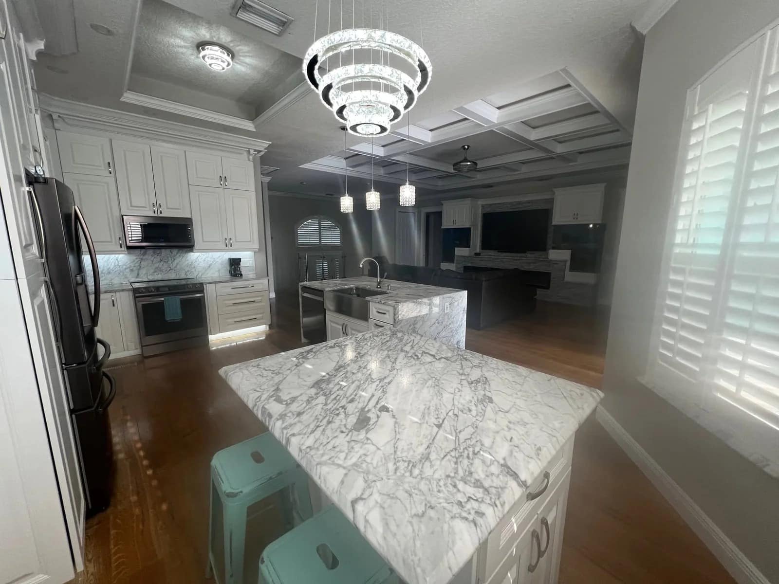 A modern kitchen with white cabinetry, stainless steel appliances, marble countertops, island seating with teal stools, pendant lights, and a large chandelier. Wooden floors and shuttered windows add light and warmth.