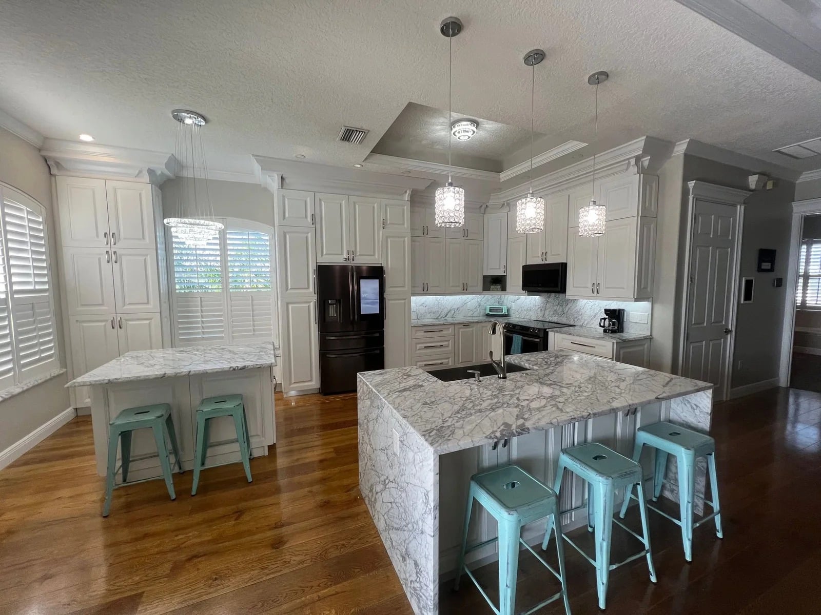 Modern kitchen with white cabinets, marble countertops, stainless steel appliances, three pendant lights, and two islands. Each island has turquoise stools. Hardwood floors and plantation shutters complete the space.