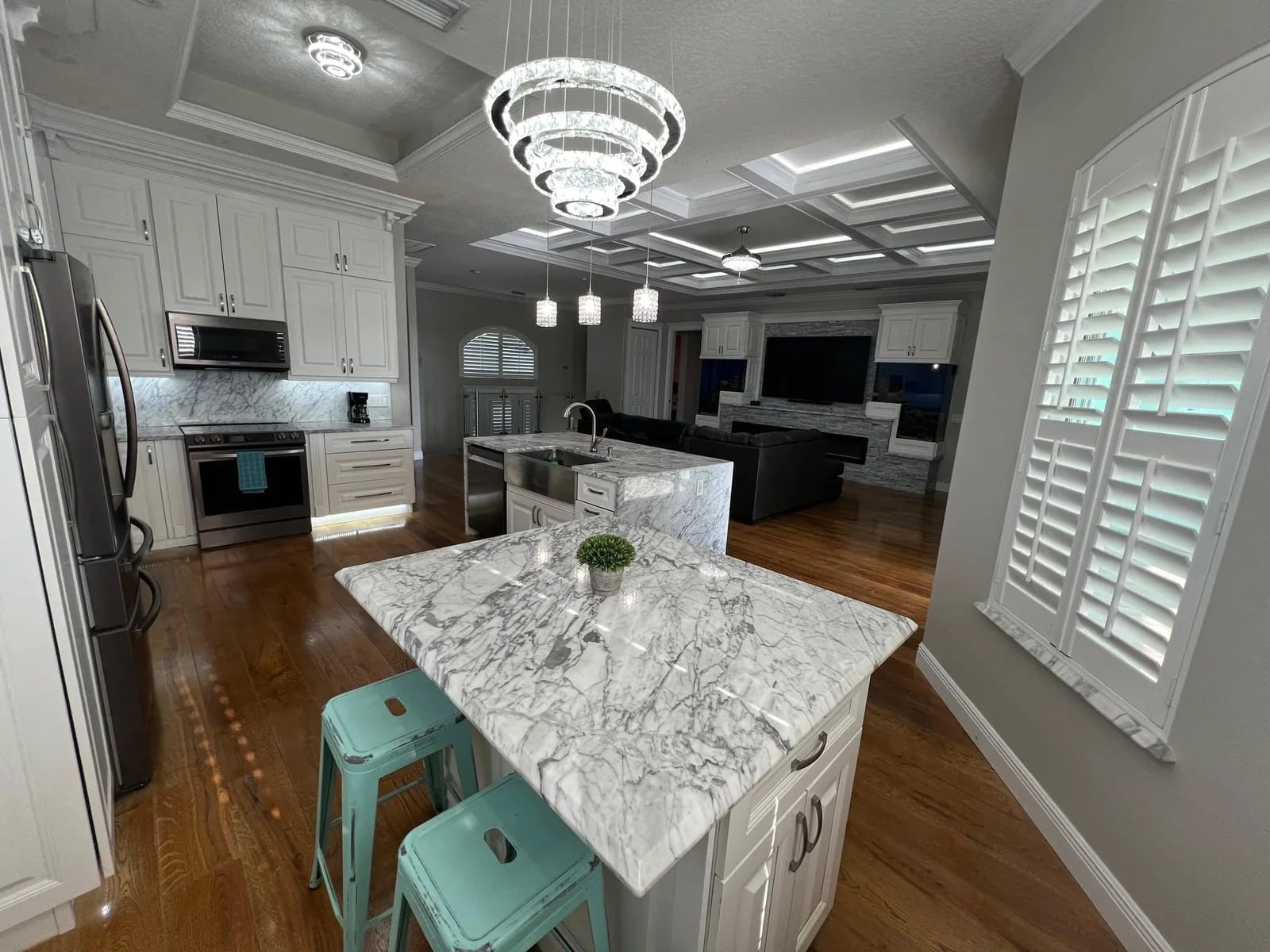 Modern kitchen with white cabinets, marble countertops, and an island with two turquoise stools. Elegant pendant lights hang above the island. Wooden floors and large windows with plantation shutters are also visible.
