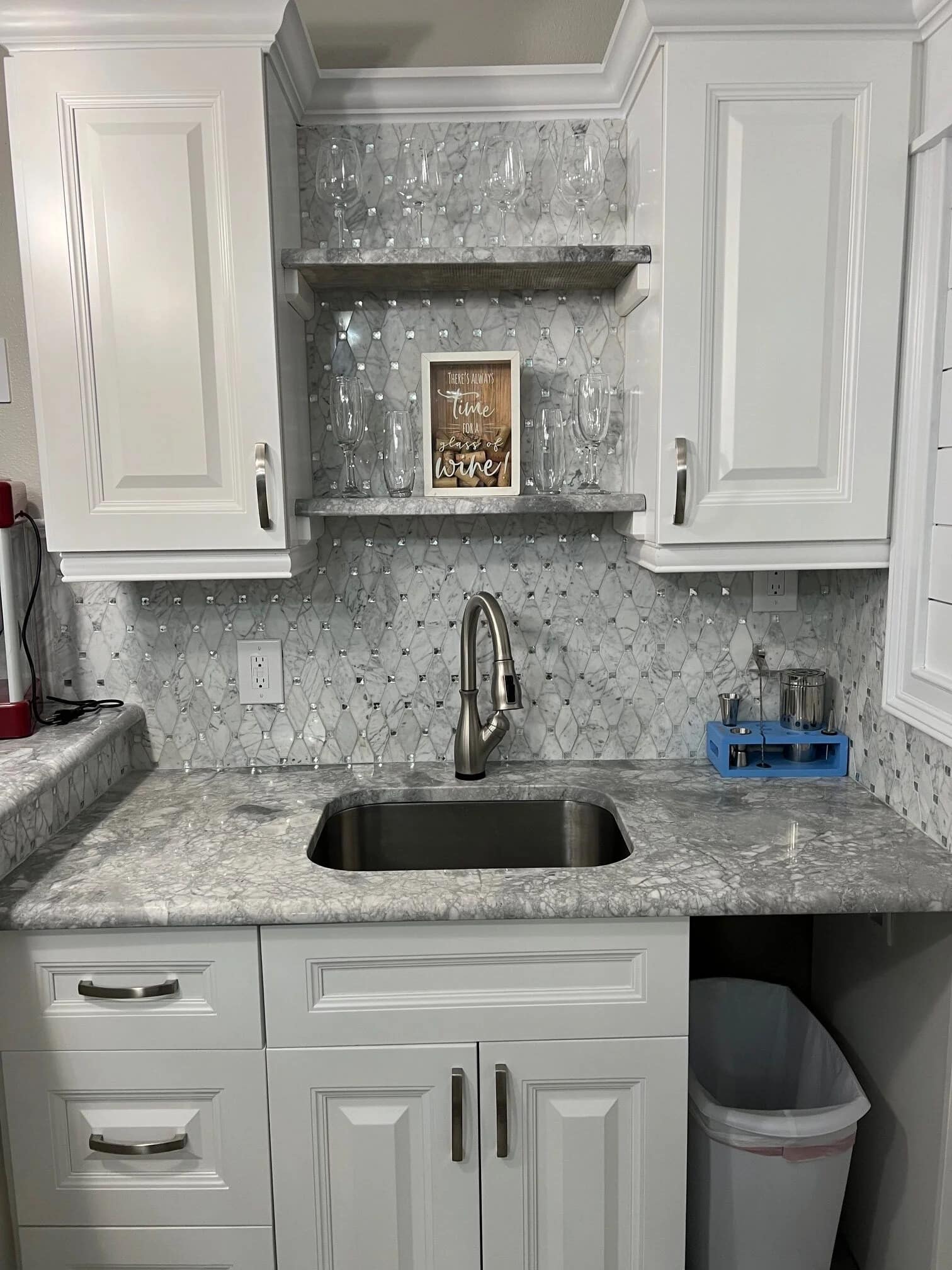 A modern kitchen sink area with white cabinets, marble backsplash, and countertop. Two shelves above the sink hold glasses and a framed sign that says “Time to drink wine.” A small blue rack holds glasses on the right.
