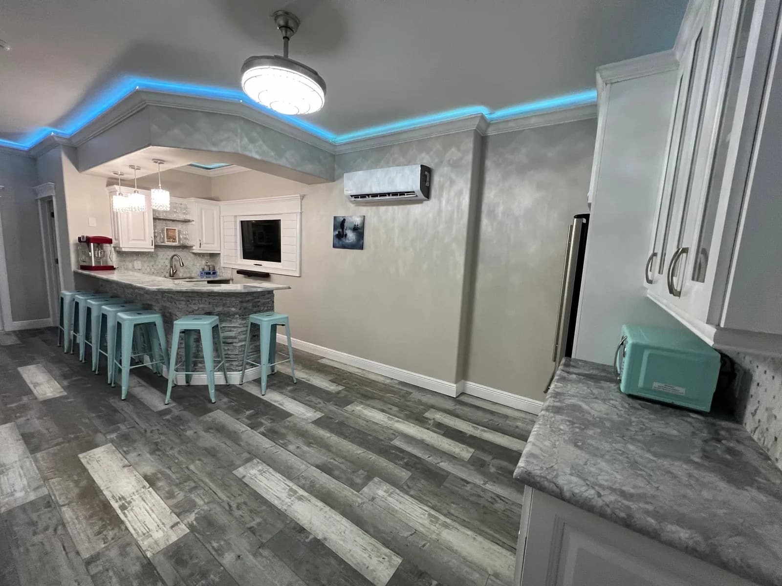 Modern kitchen with gray wood flooring, white cabinets, marble countertops, a breakfast bar with blue stools, turquoise appliances, pendant lights, and blue LED ceiling lighting. A wall-mounted air conditioner is visible.
