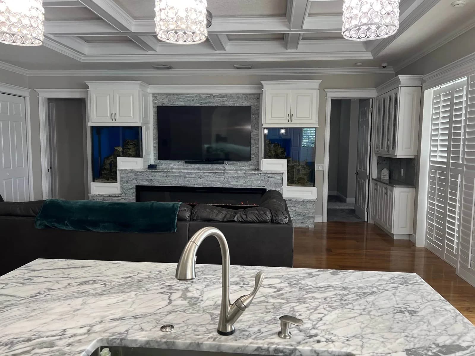 A modern living room with a marble kitchen island in the foreground, a silver sink faucet, a dark sofa, a large TV mounted on a stone accent wall, white cabinets, and wooden floors with pendant lights above.