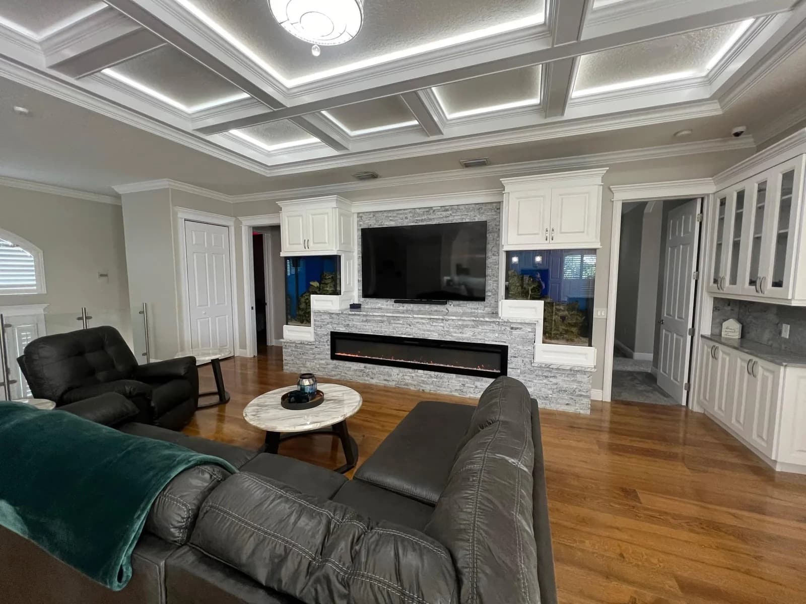 Modern living room with dark leather couches, a round coffee table, a large wall-mounted TV above a long electric fireplace, built-in white cabinetry, and hardwood floors. Coffered ceiling with recessed lighting above.