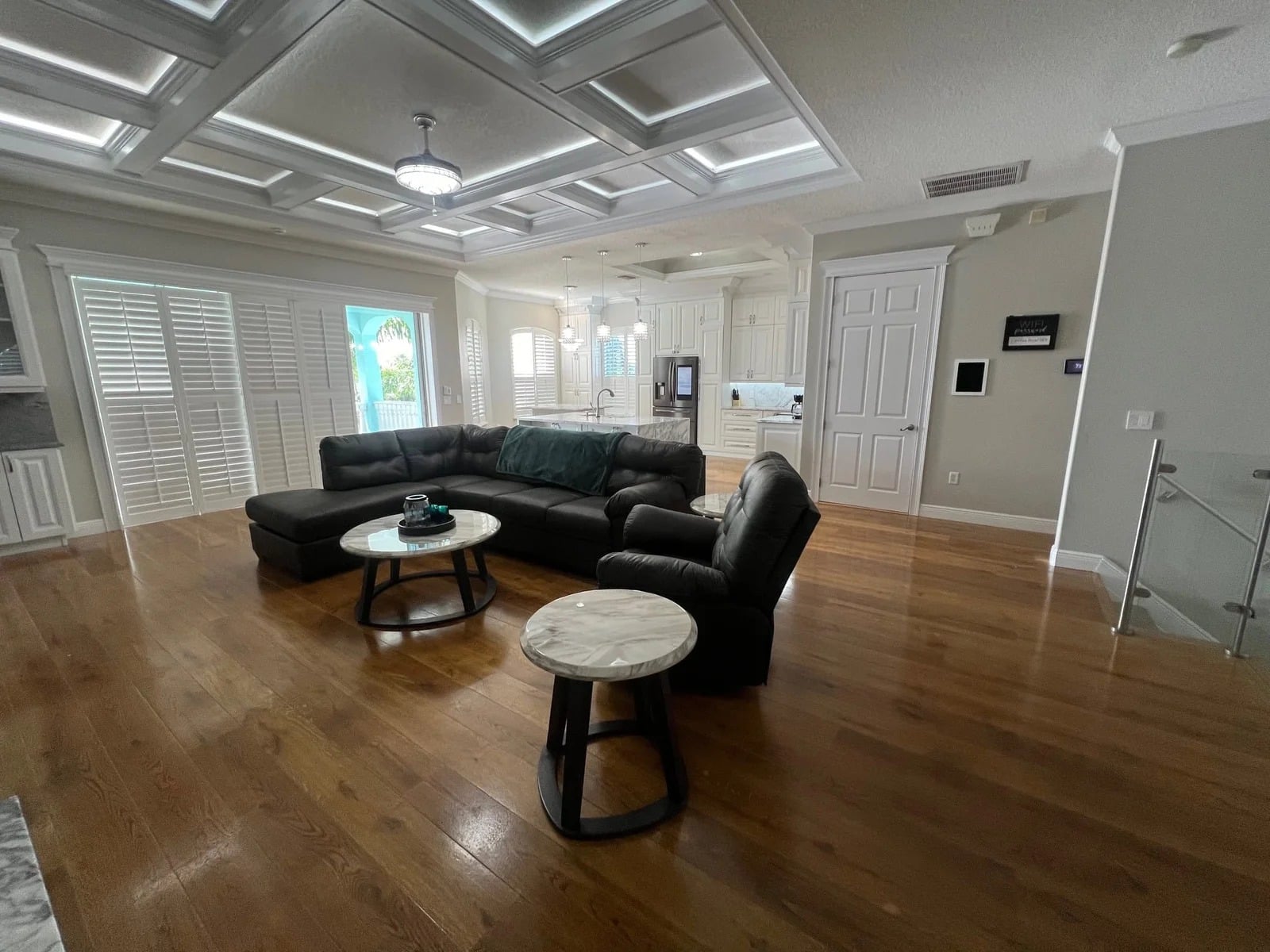 Spacious living room with wooden floors, a dark sectional sofa, two round coffee tables, and large windows with white shutters. The open layout leads to a bright, modern kitchen in the background.