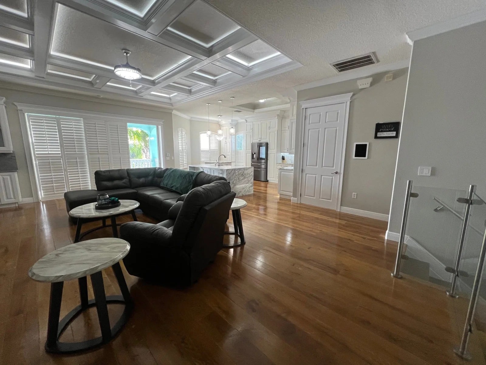 A spacious, modern living room with wooden floors, a dark sectional sofa, round marble-top tables, and an open view into a bright kitchen with white cabinets. Large windows with shutters let in natural light.