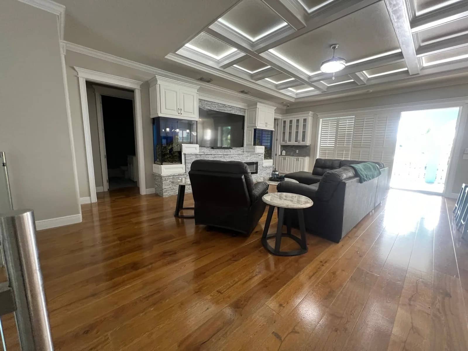 A spacious living room with polished wooden floors, a gray sofa, black armchair, white cabinets, a wall-mounted TV, coffered ceiling, and large windows with shutters letting in natural light.