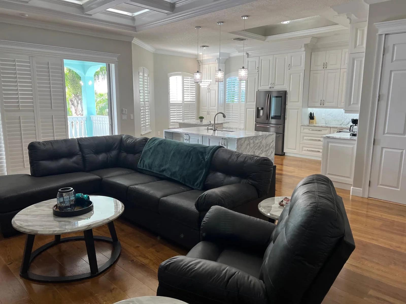 A spacious, modern living room with black leather sofas, marble-topped tables, and an open kitchen featuring white cabinetry, stainless steel appliances, and large windows letting in natural light.