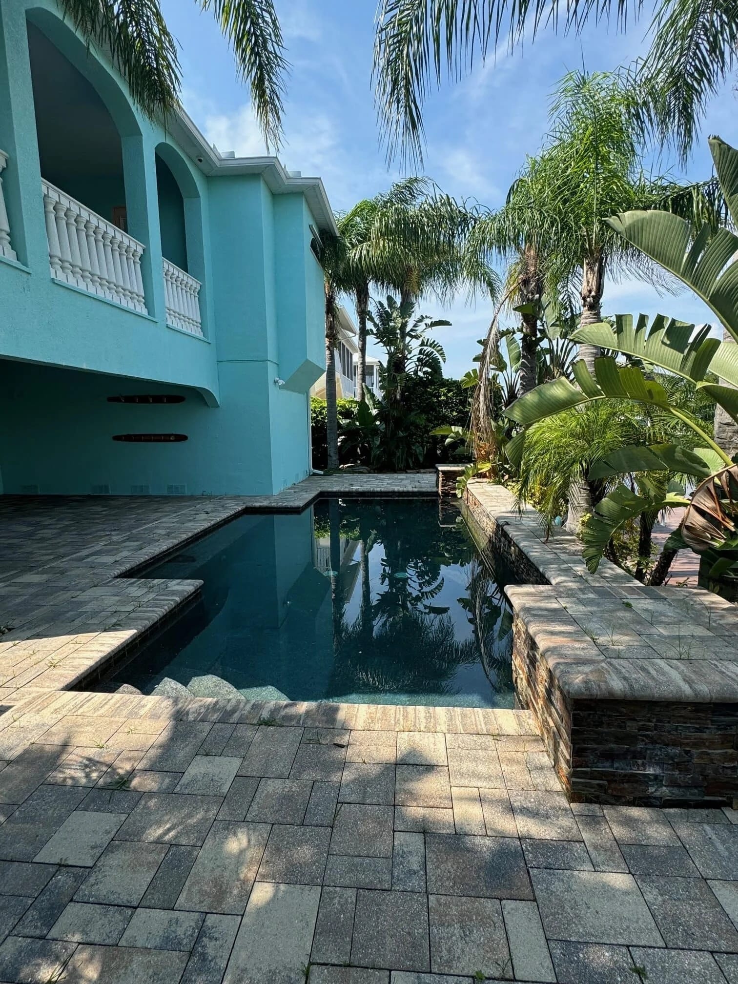 A small, rectangular backyard pool surrounded by stone tiles, tropical plants, and palm trees, next to a turquoise two-story house with white railings and arched balconies under a blue sky.