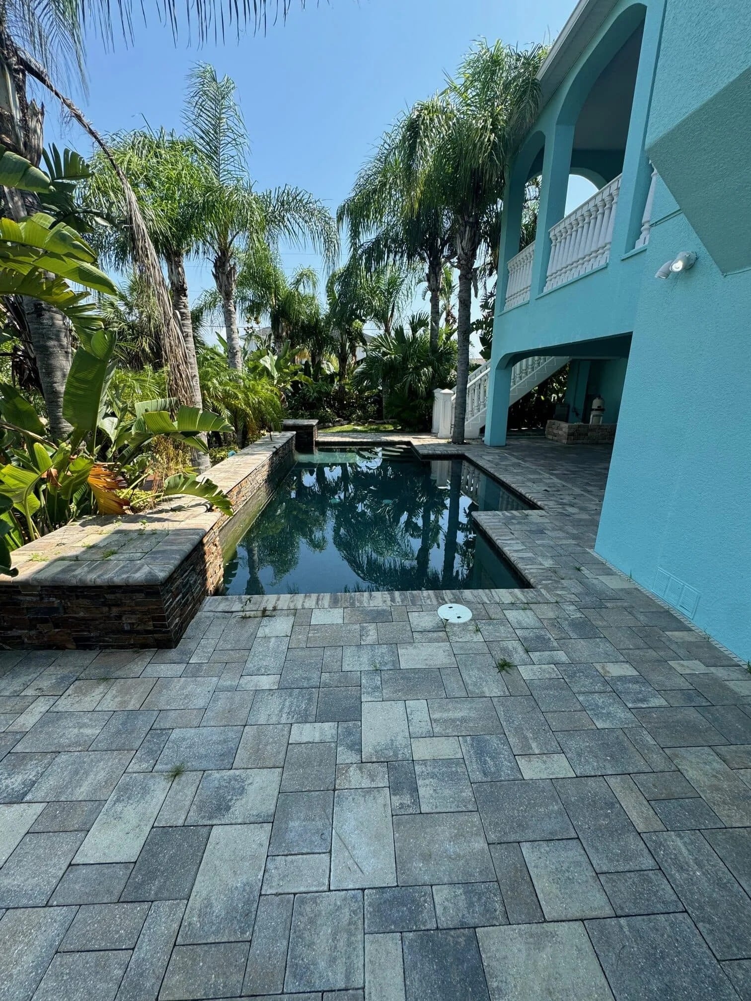 A backyard pool with a stone-tiled patio, surrounded by lush palm trees and tropical plants. The pool is beside a turquoise two-story house with an arched balcony and outdoor stairs.