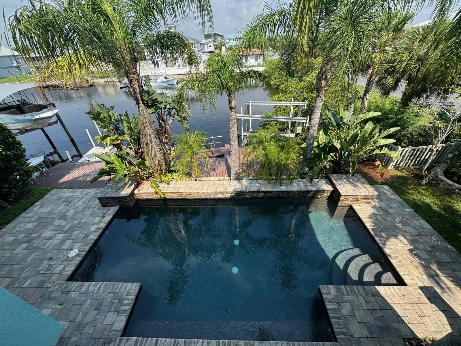 A rectangular swimming pool surrounded by palm trees and tropical plants, with a paved deck area and steps leading into the water. In the background, there is a river with docks and boats.