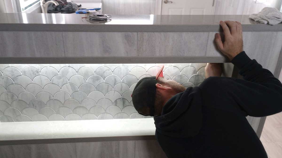 A person wearing a black hoodie and cap is installing or inspecting a tiled backsplash with a scallop pattern beneath a counter in a modern, light-colored kitchen.