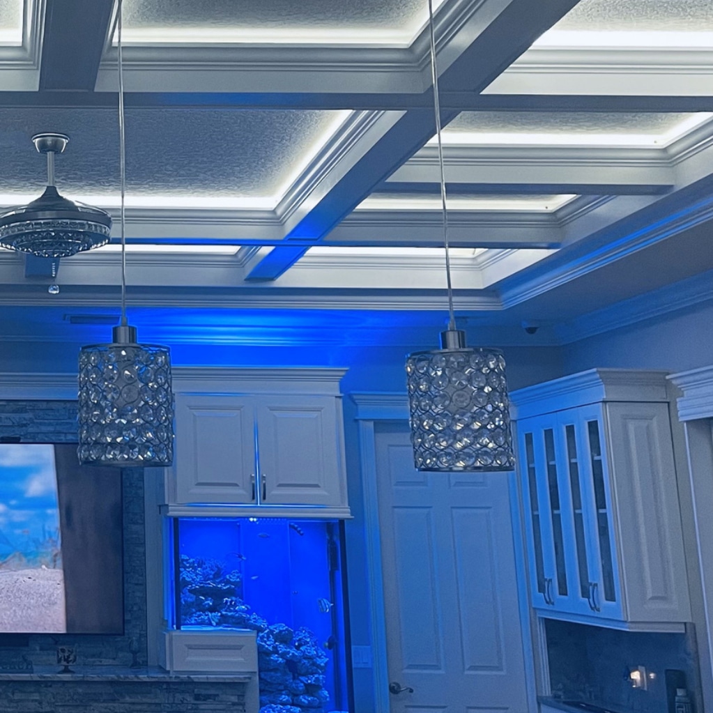 A modern kitchen with coffered ceilings lit by blue accent lights, two crystal pendant lights, white cabinets, and a large aquarium built into the wall.
