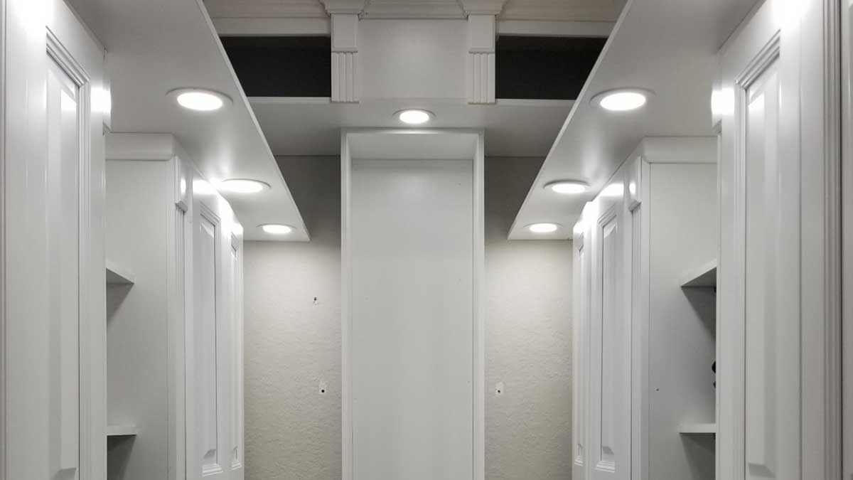A modern, empty walk-in closet with white built-in shelves and cabinets on both sides, illuminated by recessed ceiling lights. The closet is well-lit and has a clean, minimalist design.