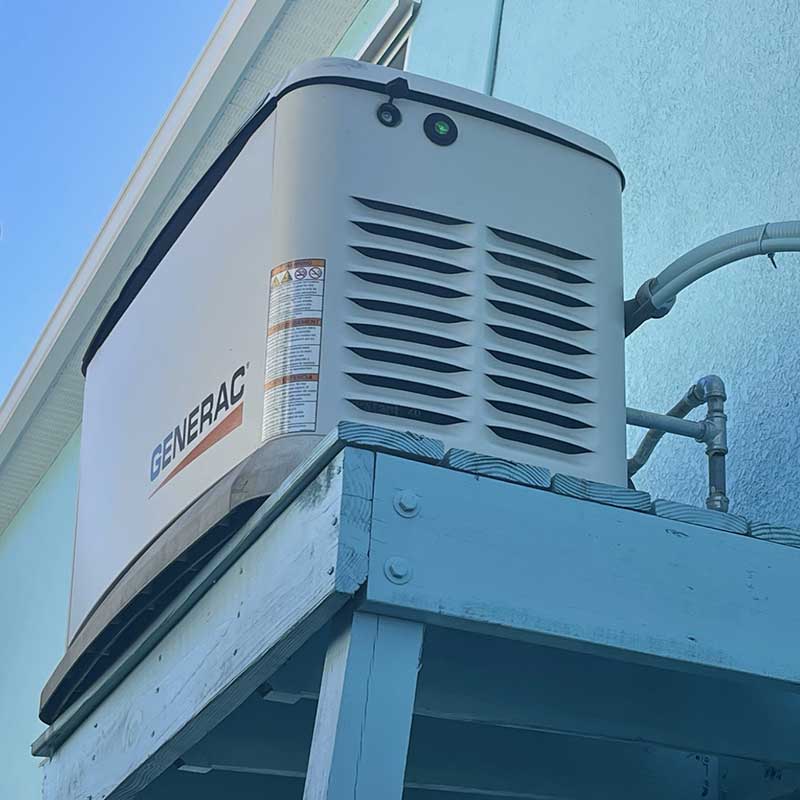 A Generac standby generator is mounted on a wooden platform attached to the side of a light blue building, with attached cables and pipes visible.