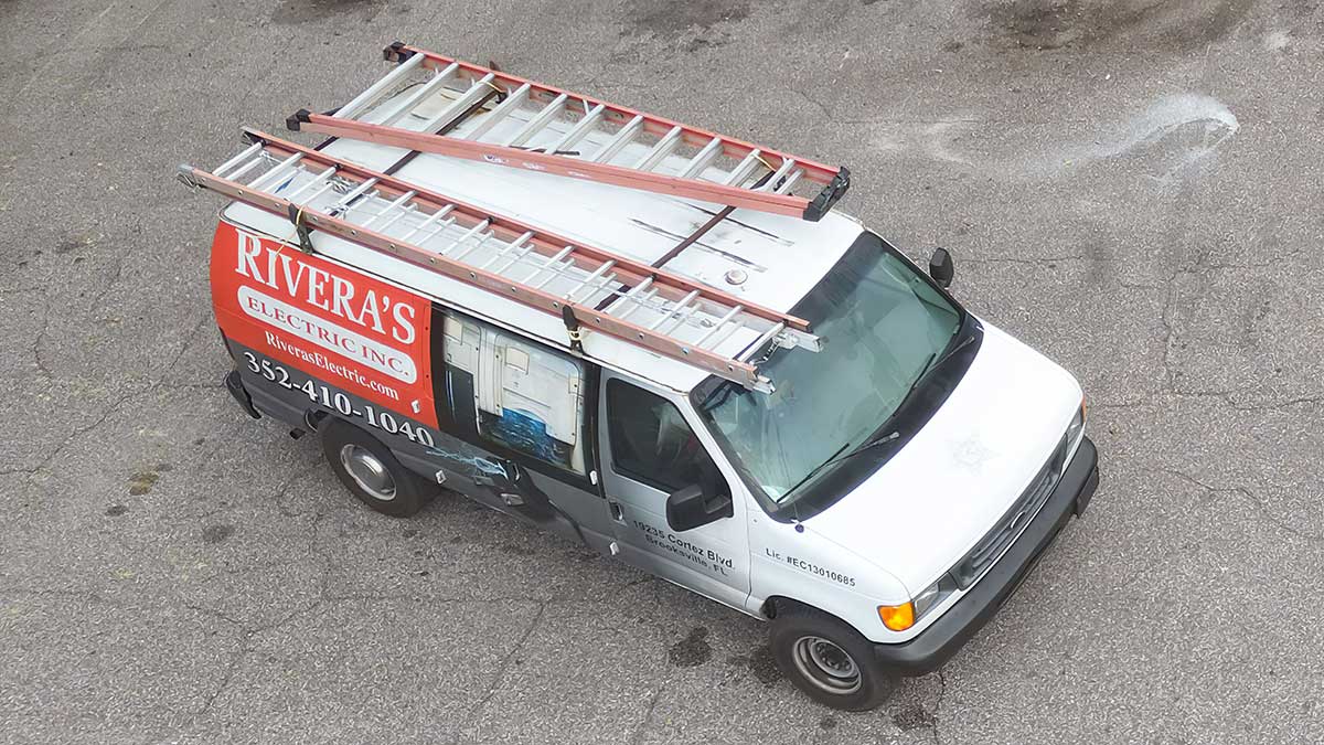 A white Rivera’s Electric Inc. service van is parked on pavement, viewed from above. The van has ladders mounted on its roof and the company’s name, logo, and phone number displayed on the side.